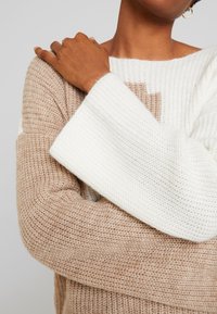 Pull en maille présentant un design bicolore avec des sections beige et blanche. Manches larges et une finition texturée qui renforcent la douceur du matériau.