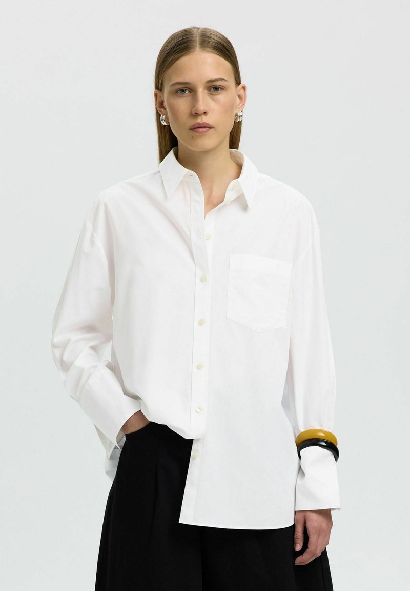 Femme aux cheveux raides porte une chemise blanche oversize boutonnée, un pantalon large noir, des bracelets dorés et noirs, ainsi que des boucles d'oreilles créoles argentées.