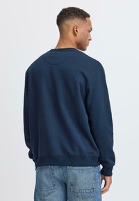 Temno modra dukserica iz mehkega bombažnega blaga, s krožnim izrezom, rebrastimi manšetami in sproščenim krojem, v kombinaciji z modrimi kavbojskimi hlačami.