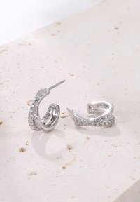 Orecchini a cerchio in argento con design intrecciato, caratterizzati da accenti scintillanti e texture liscia, posti su una superficie di pietra chiara.