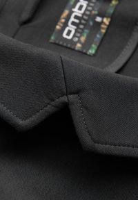 Gros plan sur un col en tissu noir avec des coutures détaillées et une étiquette de marque colorée partiellement visible à l'intérieur de l'encolure.