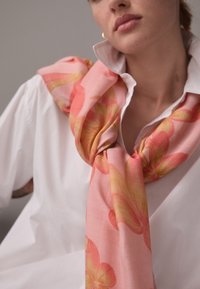 Vaaleanpunainen silkkihuivi, jossa on suuret keltaiset ja korallinpunaiset kukkakuviot, kiedottuna valkoisen napitettavan paidan kaulan ympärille. Silkkinen pinta, tyylikäs muotoilu.