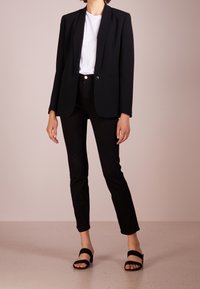 Blazer nero su misura con colletto affilato, indossato sopra una t-shirt bianca e jeans neri. Design semplice, tessuto morbido e apertura frontale. Sandali neri scivolati.