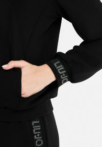 Sweatshirt noir au toucher lisse, dotée d'une poche latérale et d'un accent en cristal sur le poignet, mettant en valeur le nom de la marque.