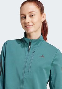 Teal athletic jacket with high collar, zipper closure, and black accents. Features the Adidas logo and a chest pocket. Smooth fabric texture.