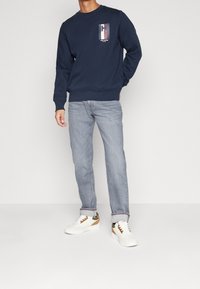 Granatowa bluza z kapturem z graficznym logo "H" na piersi, zestawiona z jasnoniebieskimi dżinsami i białymi trampkami z brązowymi akcentami.