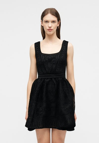 Γυναίκα με ίσια καστανά μαλλιά, φορώντας μαύρο δαντελένιο φόρεμα χωρίς μανίκια με τετράγωνο ντεκολτέ και φουντωτή φούστα, στέκεται μπροστά σε απλό φόντο.