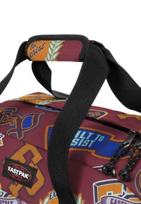 Hnědá látková taška s různými barevnými nášivkami a logy. Černé popruhy a zipy s detailními kovovými akcenty.