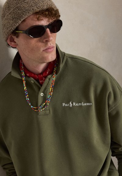 Νέος άνδρας που φοράει πουλόβερ Polo Ralph Lauren σε ελιά πράσινο, καφέ πλεκτό σκουφάκι, μαύρα γυαλιά ηλίου, κόκκινη μπαντάνα και πολύχρωμο κολιέ με χάντρες, κοιτάζοντας πλάγια.