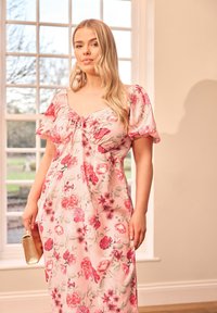 Blommig maxiklänning i rosa med korta puffärmar, en hjärtformad halsringning och en smockad framdel. Innehåller olika blommönster. Guldclutch som accessoar.
