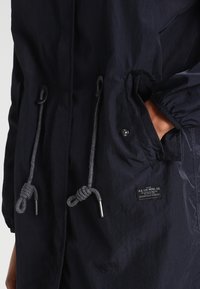 Černá bunda s předními kapsami a nastavitelnými šňůrkami, jedna ruka v kapse, viditelné logo značky poblíž kapsy.