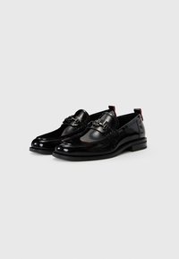Mocasines de charol negro con un diseño trenzado y un detalle decorativo de hardware plateado. Presentan una suela plana y acentos en el tacón rojo.