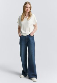 Haut blanc à manches courtes avec un motif texturé, assorti à un jean large bleu foncé et des baskets blanches. Mains dans les poches, fond neutre.