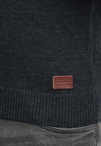 Dark gray knitted sweater with a ribbed hem, featuring a brown leather label at the bottom displaying brand and origin details.