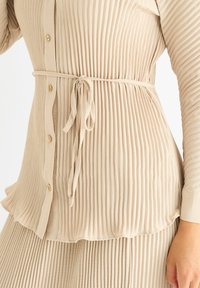 Beige pleated top with long sleeves, button closure, and a tie belt. Soft fabric and textured design enhance the overall silhouette.