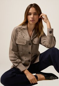 Beige Hemd mit Knöpfen, dekorativen Nieten, zwei Brusttaschen und langen Ärmeln. Kombiniert mit marineblauen Hosen und schwarzen Slippers mit Nieten.