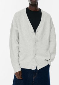 Cardigan a maglia grigia chiaro con scollatura a V, bordi a coste e cinque bottoni, indossato sopra una t-shirt nera. Il tessuto appare morbido e texturizzato.