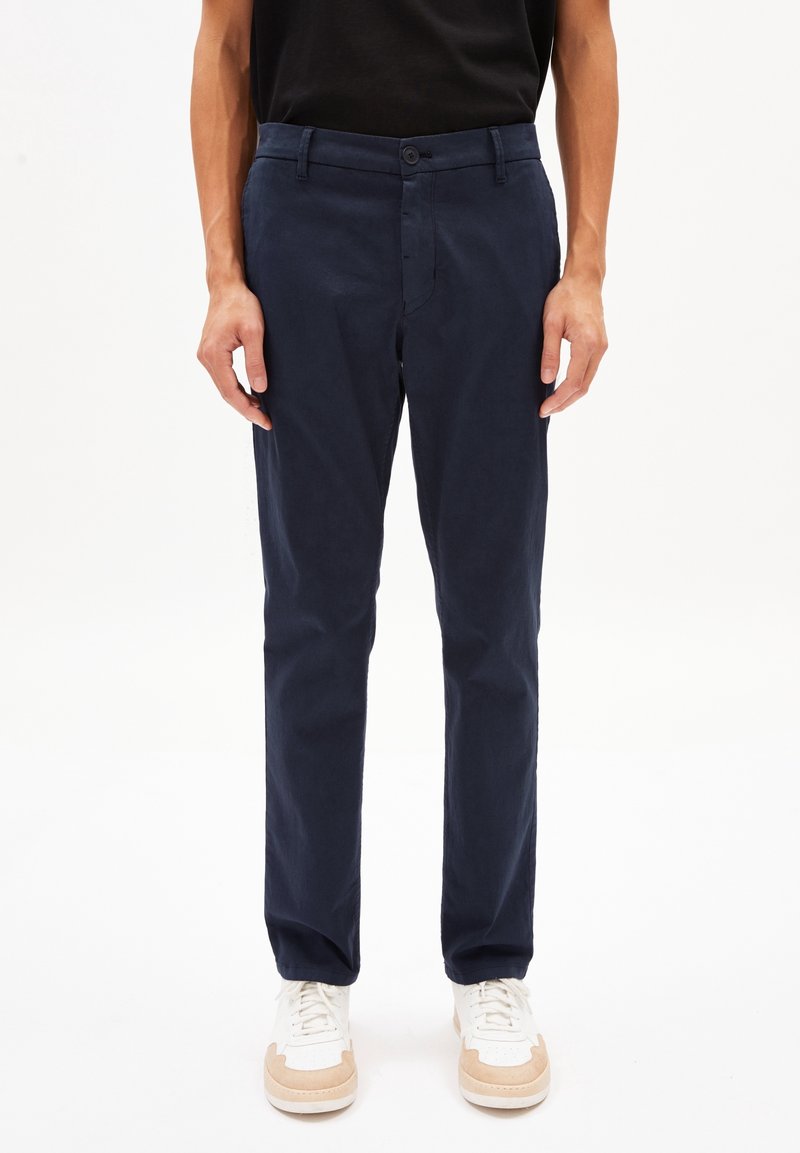 Muž v tmavě modrých rovně střižených kalhotách a bílých teniskách stojící na jednobarevném bílém pozadí.