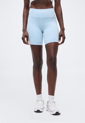 Persona con shorts de ciclismo de tiro alto azul claro y zapatos deportivos blancos con calcetines blancos, de pie contra un fondo liso y claro.