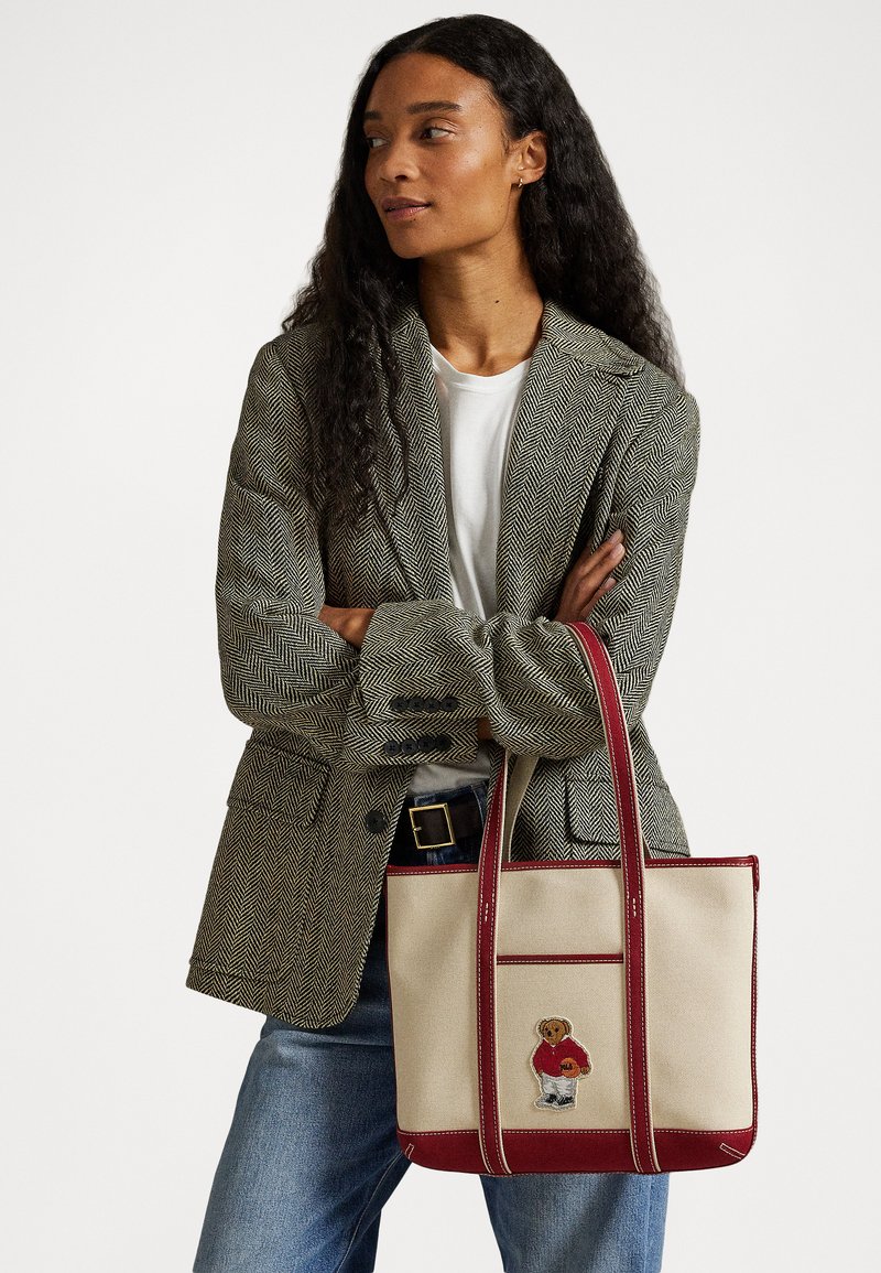 Tote bag gemaakt van crèmekleurig canvas met rode accenten en handvatten. Heeft een geborduurd berenontwerp in een rode trui en blauwe broek.