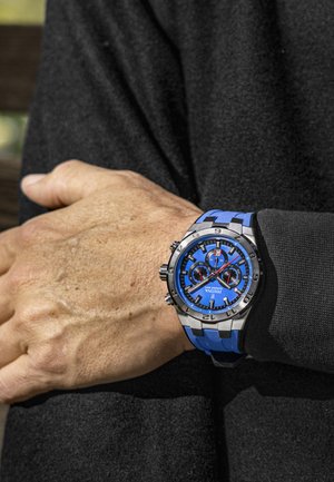 Montre de sport bleue et argentée au poignet. Elle présente un cadran rond, de grands chiffres, deux sous-cadrans et un bracelet en silicone à motifs.