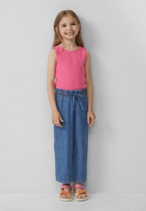 Een jong meisje staat en glimlacht, gekleed in een roze mouwloos topje, hoge blauwe denim broek en kleurrijke sandalen.