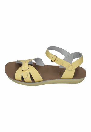 Yellow leather open-toe sandal with ankle and front buckle straps on a brown flat sole against white background.