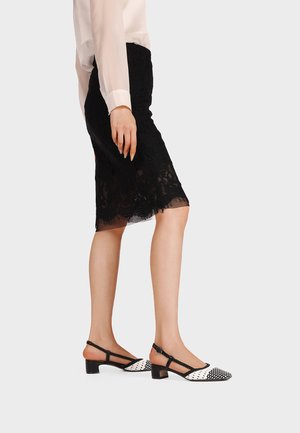 Donna con gonna nera in pizzo fino al ginocchio, camicetta bianca trasparente e scarpe slingback bianche e nere con tacco basso, in piedi di lato.