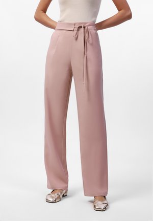 Femme portant un pantalon large taille haute rose poudré avec une fine ceinture à nouer et des chaussures plates métalliques à bout carré.