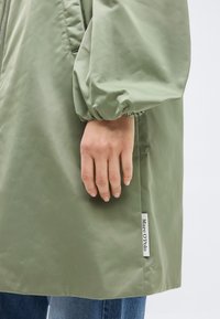 Kéz pihen az olívazöld kabát ujjánál, amelyen elasztikus mandzsetta van; a "Marc O'Polo" címke látható a kabát szegélyén, a kék farmert felül.