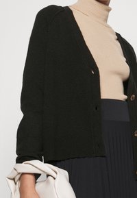 Czarny kardigan z dekoltem w serek, o fakturze prążkowanej i zapięciami na guziki. Noszony na beżowym, prążkowanym golfie, z jasną torebką.