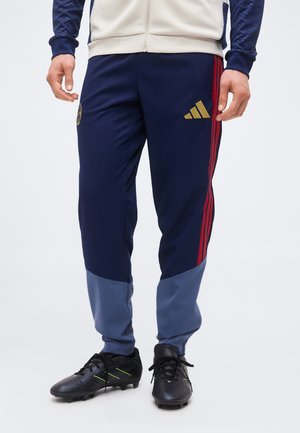 Mann trägt marineblaue und graue Sporthose mit roten Streifen und schwarzen Stollen, steht vor einem schlichten weißen Hintergrund.