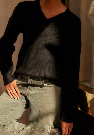 Pull en maille côtelée noire avec col en V, associé à un jean en denim bleu clair. Le tissu semble doux, avec une coupe décontractée et des épaules tombantes.
