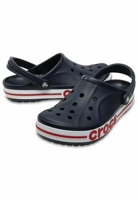 Navy blue Crocs clogs with ventilation holes, heel strap, and white/red striped sole; features a prominent "Crocs" logo on the side.