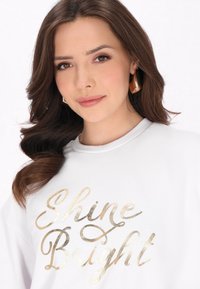 Witte sweatshirt met "Shine Bright" in goud metallic schrift. Het model heeft middelbruin haar en draagt grote gouden ringoorbellen.