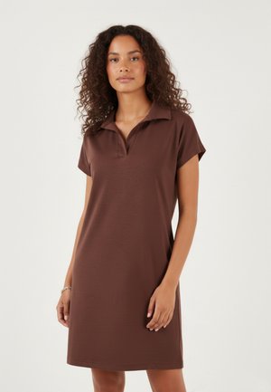 Kvinde med krøllet hår iført en brun polokjole med korte ærmer og knælængde stående mod en ensfarvet lyse baggrund.