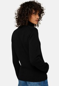 Femme aux cheveux bouclés portant un pull noir à col roulé et un jean bleu, se retournant contre un fond uni.