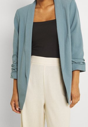 Vrouw draagt een lichtblauwe open blazer met geplooide mouwen, een zwart mouwloos topje en een crèmekleurige broek met hoge taille tegen een effen achtergrond.