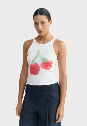 Femme aux cheveux coiffés en arrière portant un haut blanc sans manches orné d'un motif de cerises rouges et vertes, associé à un pantalon taille haute foncé.