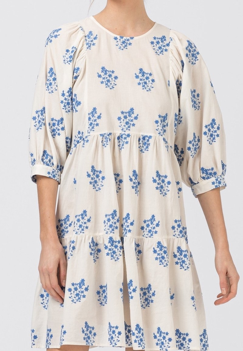 Robe à manches courtes couleur crème avec motif floral bleu, corsage froncé et jupe à étages. Réalisée en tissu léger à la texture lisse.