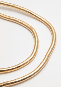 Questa catena in oro metallico presenta un design liscio e arrotondato, con una superficie strutturata e rigata, e una forma flessibile, posata su uno sfondo bianco.