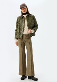 Gestepptes olivgrünes Jacket mit braunem Kragen, cremefarbenem Pullover und karierten Schlaghosen. Eine braune Lederkappe und klobige schwarze Stiefel runden das Outfit ab.