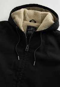Giacca nera con zip, con una fodera in pile texturizzato color crema. Presenta un cappuccio regolabile e un'etichetta interna con le istruzioni per la cura.
