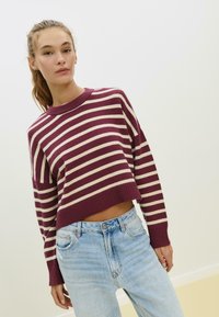 Burgunderfarbener und cremefarbener gestreifter Pullover mit geripptem Rundhalsausschnitt und langen Ärmeln, kombiniert mit hellblauen, verwaschenen Jeans.