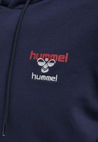 Marineblauwe hoodie met witte en rode geborduurde "hummel" logo's. Heeft een trekkoord capuchon en een textuurstof met een gladde afwerking.