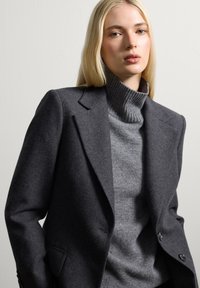 Blazer de lana gris con hombros estructurados y cierre de un solo botón, superpuesto a un suéter de cuello alto acanalado gris claro. Textura suave.