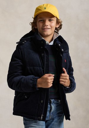 Junge mit lockigem Haar trägt eine gelbe Kappe, eine marineblaue Steppjacke, einen dunklen Pullover und hellblaue Jeans und lächelt vor einem einfarbigen Hintergrund.