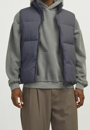 Personne portant un sweat à capuche gris, un gilet matelassé bleu marine et un pantalon marron, debout les bras détendus sur un fond uni clair.