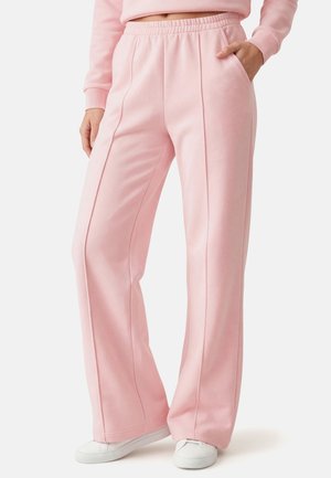Kobieta nosząca jasnoróżowe szerokie spodnie z detalami szwów, elastycznym paskiem, kieszeniami bocznymi, połączone z białymi tenisówkami i dopasowanym różowym topem.