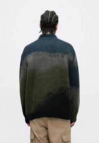 Πλεκτό πουλόβερ με σχεδίαση με διαβαθμίσεις σε σκούρο πράσινο, γκρι και ναυτικό μπλε χρώμα. Μαλακή υφή, ψηλός λαιμός και χαλαρή εφαρμογή.